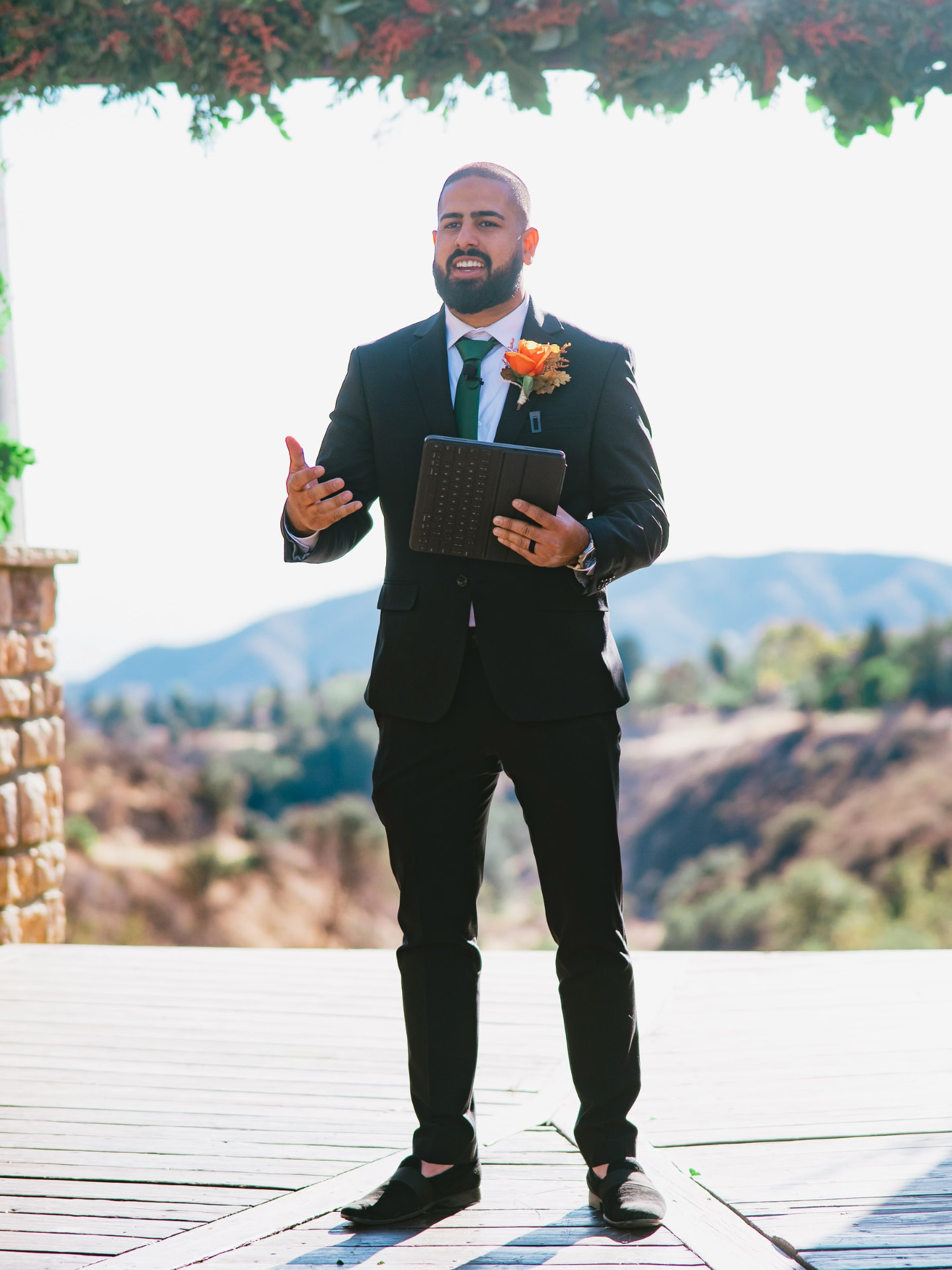 Matt Perry in a black suit and green tie speaking at an outdoor ceremony, holding a folio with hills behind him