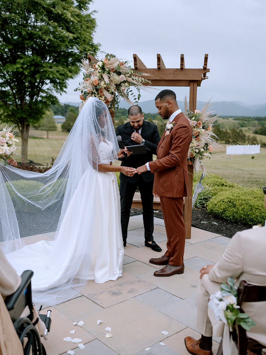 A bride in a long veil and a groom in a rust suit exchange rings under a floral arch while Matt Perry reads beside them