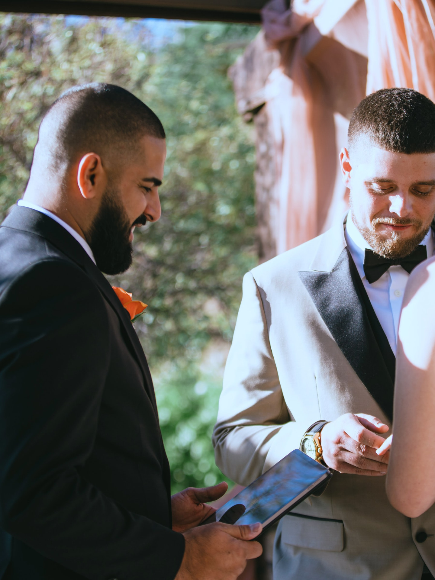 Matt Perry reading from a folio beside a groom in a light grey tuxedo placing a ring on his partner's finger